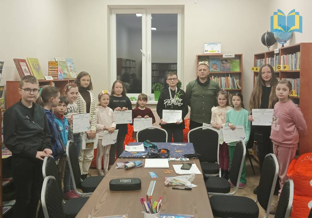 Zdjęcie: Dzieci stoją w bibliotece w półkolu. Niektóre z nich trzymają dyplomy za najlepsze komiksy o Niegowie. Przed nimi znajduje się duży stół z przyborami biurowymi (kartki, kredki).